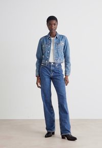 Denim dzseki két elülső zsebbel, fehér felsőre húzva, magas derekú kék farmernadrággal párosítva. A modell semleges háttér előtt áll.