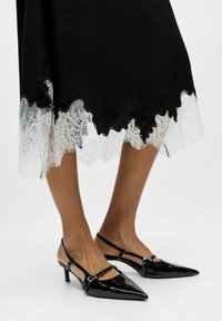 Naine kannab musti läikivaid terava ninaga slingback-kingi keskmise kõrgusega kontsadega ning musta seelikut, millel on valge pitsäär.