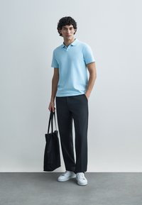 Hellblaues Poloshirt mit kurzen Ärmeln, kombiniert mit dunkelgrauer Hose und weißen Turnschuhen. Schwarze Tragetasche in der linken Hand gehalten.