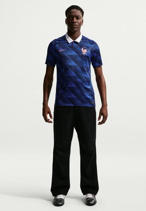 Mand står vendt fremad iført blå mønstret Frankrigs fodboldtrøje, sorte bukser og sorte-hvide sko mod hvid baggrund.