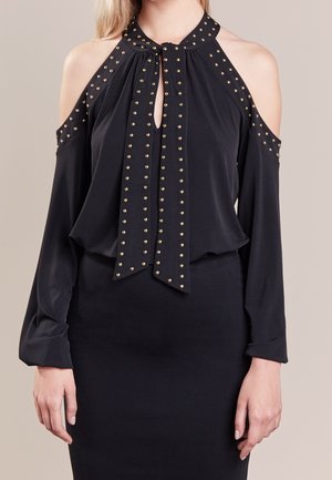 Schwarze Cold-Shoulder-Bluse mit goldenen Nieten, die Schlüssellöcher und Knotenakzente umrahmen, kombiniert mit einem passenden schwarzen Rock.