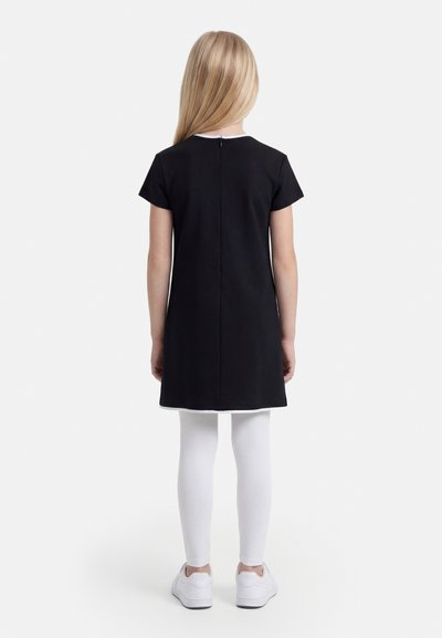 Child with long blonde hair wearing a black short-sleeve dress, white leggings, and white sneakers, standing facing away.