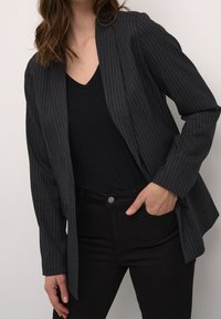 Zwarte blazer met krijtjestrepen en een getailleerde pasvorm, open voorkant, lange mouwen en een licht gestructureerde stof, gecombineerd met een zwart V-hals top en zwarte broeken.