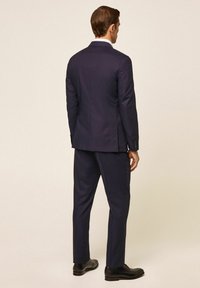 Mornarskoplavi odijelo s krojenim krojem, s jednim prorezom i glatkom teksturom. Upareno s crnim cipelama, prikazano s leđa.