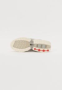 Podeszwa butów sportowych z teksturowanej gumy, przezroczystymi sekcjami i czerwonymi okrągłymi akcentami po bokach, głównie w białych i szarych odcieniach.