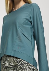 Tealfarbens langärmeliges Athletikoberteil mit genähten Details, strukturiertem Seiteneinsatz und dezentem Logo. Kombiniert mit Leggings im Animal-Print.