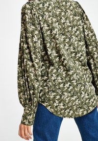 Blusa verde oliva con stampa floreale bianca, maniche lunghe a sbuffo arricciate ai polsini, indossata con jeans di denim blu.