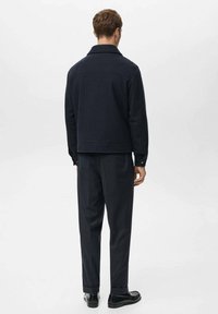 Granatowa kurtka z mieszanki wełny z kołnierzem, detalem na karku i mankietami na guziki, zestawiona z ciemnymi spodniami i butami.