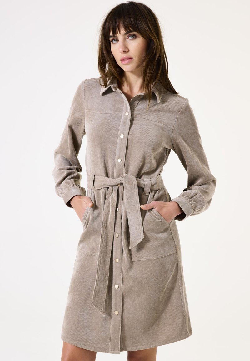 Vestido camisero de pana beige con mangas largas, cuello, botones frontales, cinturón de lazo y dos bolsillos laterales. Textura suave, diseño hasta la rodilla.