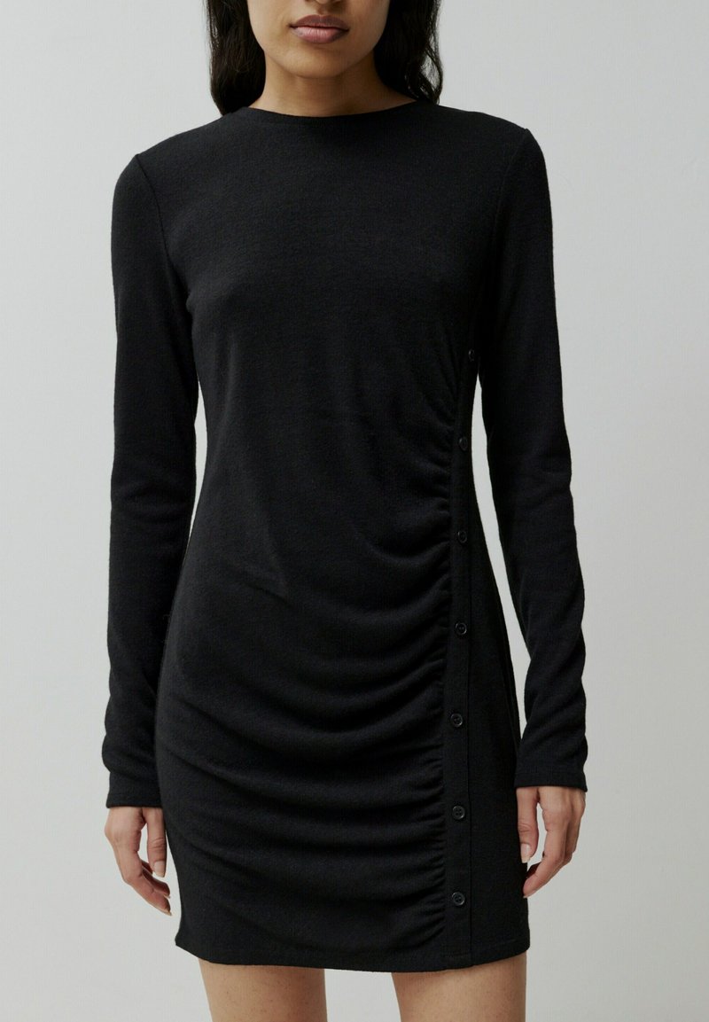 Schwarzes langärmliges Kleid mit seitlichen Rüschen und Knöpfen. Glattes Material mit taillierter Passform und Rundhalsausschnitt. Einfacher, minimalistische Stil.