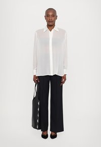 Transparent white blouse with a classic collar, button-down front, and long sleeves, paired with black wide-leg trousers and a black tote bag.