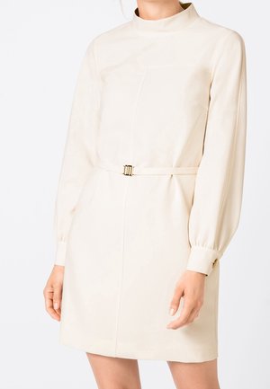 Femme portant une robe crème à manches longues avec un col montant et une ceinture fine à la taille avec une boucle dorée.