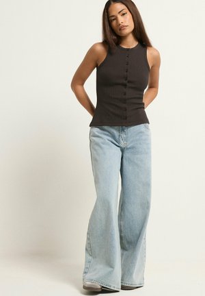 Femme debout, les mains dans le dos, portant un haut noir sans manches boutonné et un jean large bleu clair sur fond neutre.