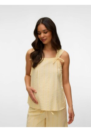 Donna incinta con capelli lunghi e scuri che indossa una canotta gialla e pantaloni abbinati, tenendo dolcemente la pancia e sorridendo verso il basso.