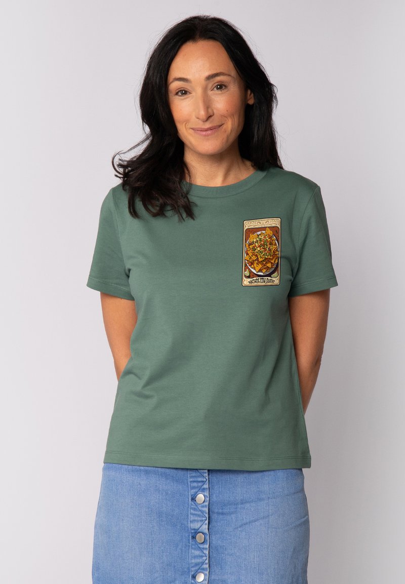 T-shirt en coton vert avec un col rond, présentant un patch graphique coloré à l'avant. Porté avec une jupe en denim bleu clair.