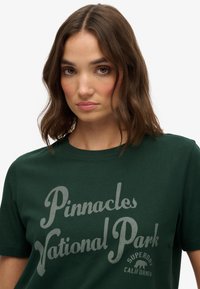 Mørkegrønt bomulds t-shirt med et grafisk tryk af "Pinnacles National Park" i lysegrå, med mærkets navn og logo.