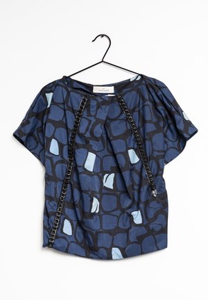Blouse bleue et noire à manches courtes avec des motifs abstraits en carrés et un détail de bordure noire, suspendue à un cintre en fil de fer noir.