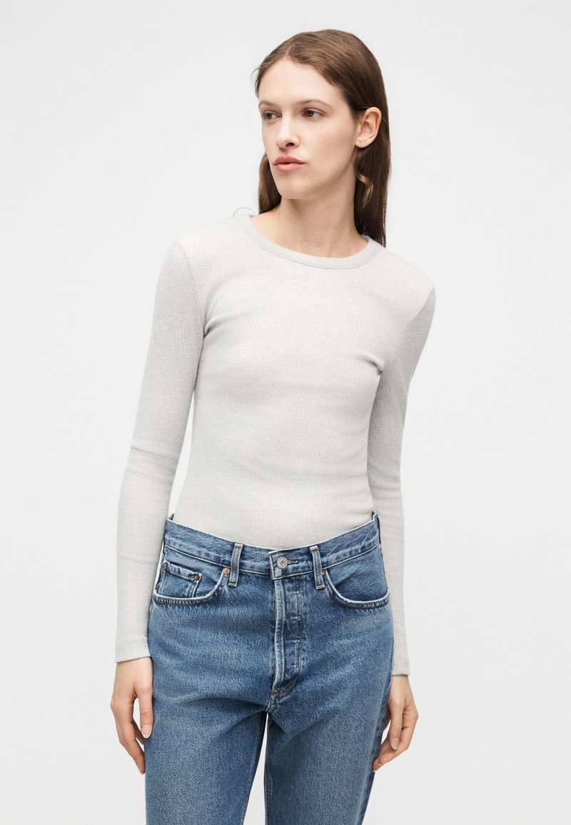 Femme portant un haut à manches longues côtelé gris clair rentré dans un jean bleu taille haute, debout devant un fond blanc uni.