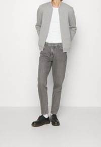 Veste bomber grise sur un t-shirt blanc, associée à un jean gris clair et des chaussures noires à lacets, sur un fond blanc uni.