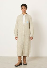 Bež pletena jakna z visokim ovratnikom, dolgimi rokavi, zapiranjem na gumb in nalepko. Model jo kombinira z rjavimi gležnjarji.