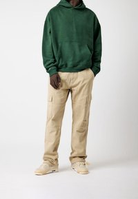 Grüner Kapuzenpullover mit einer Fronttasche, kombiniert mit beigen Cargo-Hosen mit seitlichen Taschen. Die Schuhe sind beige Sneaker.