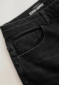 Jean en denim noir en gros plan montrant la poche avant, la boucle de ceinture et l'étiquette intérieure de la ceinture indiquant "Next Denim Supply."