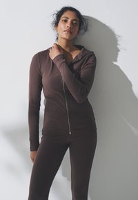 Felpa marrone a costine con zip e maniche lunghe, abbinata a pantaloni aderenti coordinati. Texture liscia e design slim fit, posata contro una parete bianca.