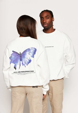 Zwei Personen tragen weiße Sweatshirts und beige Hosen; eine schaut nach vorne, die andere zeigt einen großen blauen Schmetterling und den Text „Soul and Resurrection“ auf dem Rücken.