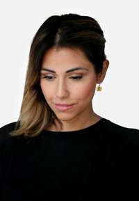 Gylne hoop øredobber med en glatt, avrundet design. Modellen har på seg en svart topp og har mellomlang, bølgete brunt hår.
