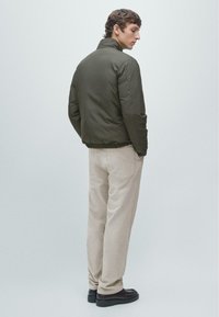 Giacca imbottita verde oliva con collo alto e orlo elasticizzato, abbinata a pantaloni beige chiaro e scarpe nere. Design semplice.