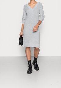 Femme portant une robe en maille côtelée grise à col en V, des bottines noires et tenant une pochette noire, marchant sur un fond uni.