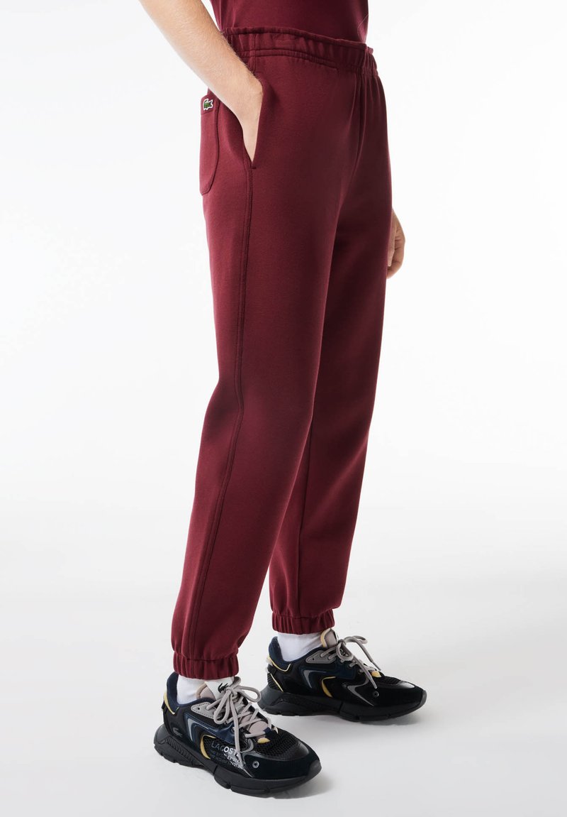 Pantaloni jogger bordeaux con cintura e polsini elastici, tasche laterali, realizzati in tessuto morbido. Abbinati a scarpe da ginnastica nere.