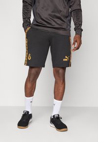 Shorts noirs avec des accents et motifs orange, portant un logo et le texte "KING". Associés avec des chaussures noires et des chaussettes de sport blanches.