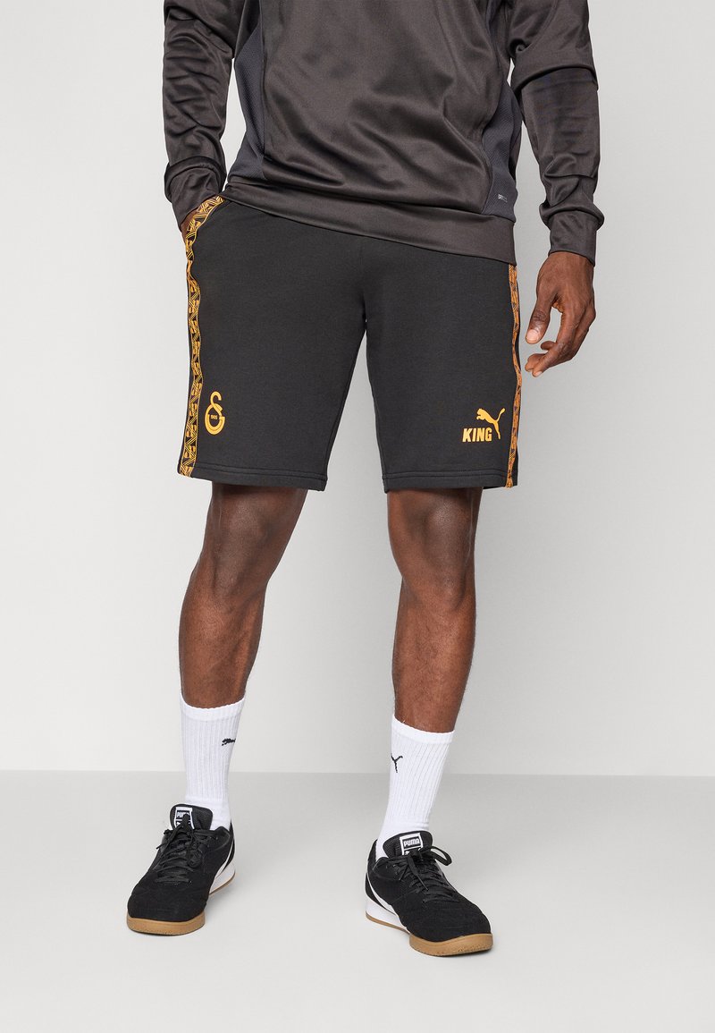 Shorts noirs avec des accents et motifs orange, portant un logo et le texte "KING". Associés avec des chaussures noires et des chaussettes de sport blanches.
