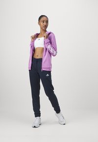 Fialová mikina na zips s bielym logom, čierne elastické nohavice a biele tenisky. Oblečenie obsahuje priliehavú bielu športovú podprsenku pod ním.