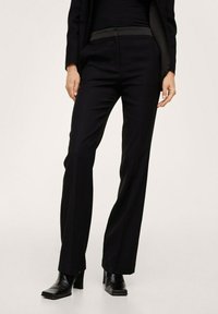 Pantalon noir sur mesure avec une taille haute, des jambes droites et une texture de tissu lisse, associé à des bottines en cuir noir.