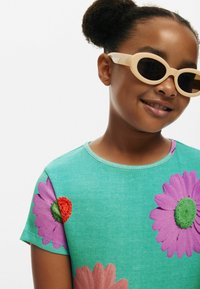 Tyrkysové bavlnené tričko s veľkými fialovými a červenými kvetinovými potlačami, s rebrovanou textúrou a okrúhlym výstrihom. Doplnilo s svetlými slnečnými okuliarmi.