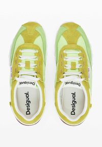 Zelené a žluté semišové tenisky s bílými tkaničkami, polstrovaným límcem a logem. Obsahují kontrastní textury a bílou gumovou podrážku.