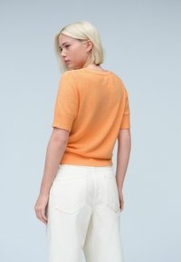 Maglione arancione a maniche corte con un design a maglia testurizzata, colletto rotondo e orlo a coste, abbinato a pantaloni bianchi a gamba larga.