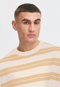 Ljust beige randig t-shirt med horisontella ränder i mjukt brunbeige, rund halsringning, korta ärmar och slät bomullstextur.