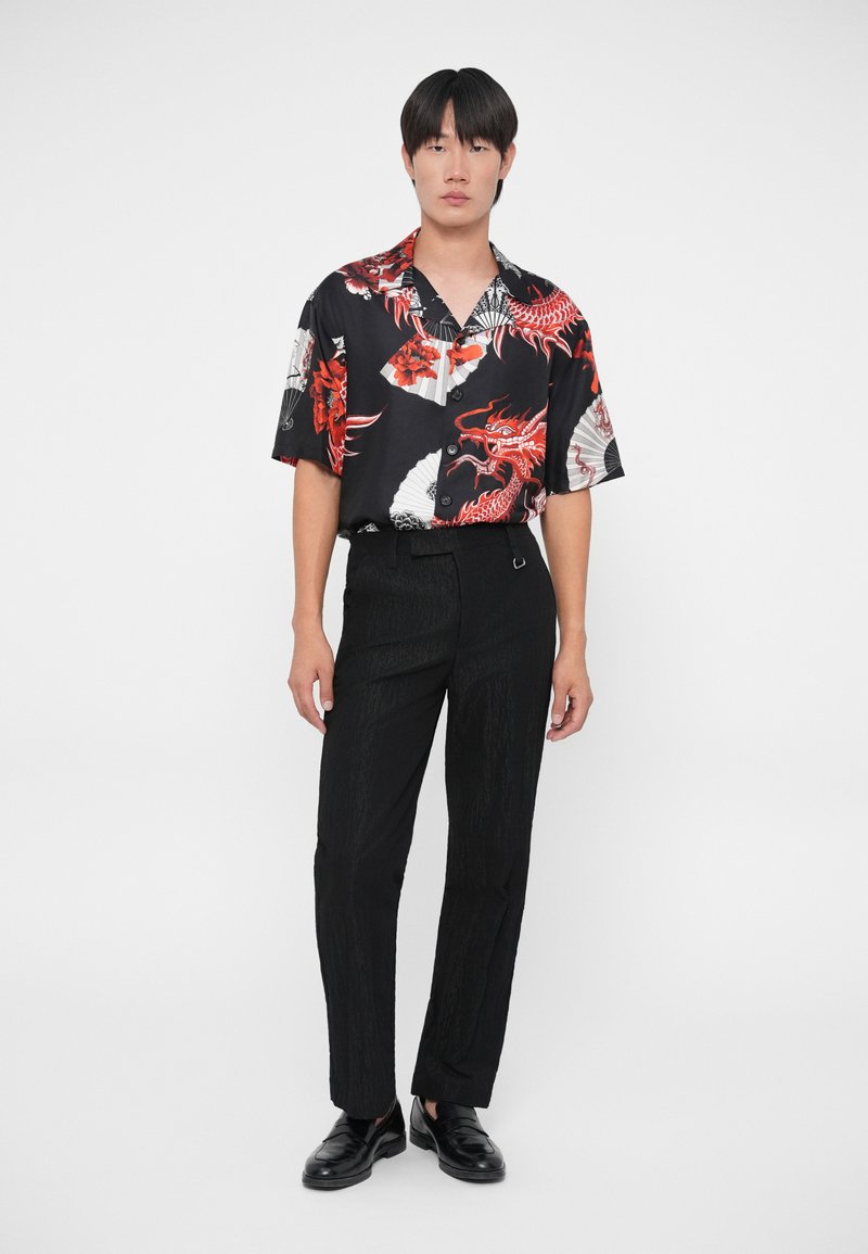 Schwarzes kurzärmeliges Hemd mit rotigem Drachen- und Fächer-Muster, kombiniert mit schwarzen strukturierten Hosen. Schwarze Schuhe runden das Outfit ab.