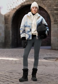 Kurtka polarowa z niebiesko-beżowym abstrakcyjnym wzorem, biała bluza z kapturem, szare legginsy oraz czarne zimowe buty z teksturowanym mankietem, trzymająca czarną torbę.