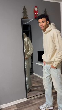 Beige hoodie and light blue jeans, standing next to a tall mirror. Room features gray walls and wooden flooring. Minimalistic decor.