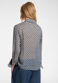 Camicia trasparente con un motivo a foglia blu e beige, polsini con bottoni e colletto. Ha una vestibilità morbida ed è abbinata a pantaloni di denim.