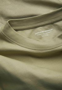 Camiseta de algodón color verde oliva con cuello redondo acanalado. Presenta una etiqueta con "JACK & JONES ORIGINALS", tamaño "Grande" y origen "Hecho en Bangladesh."