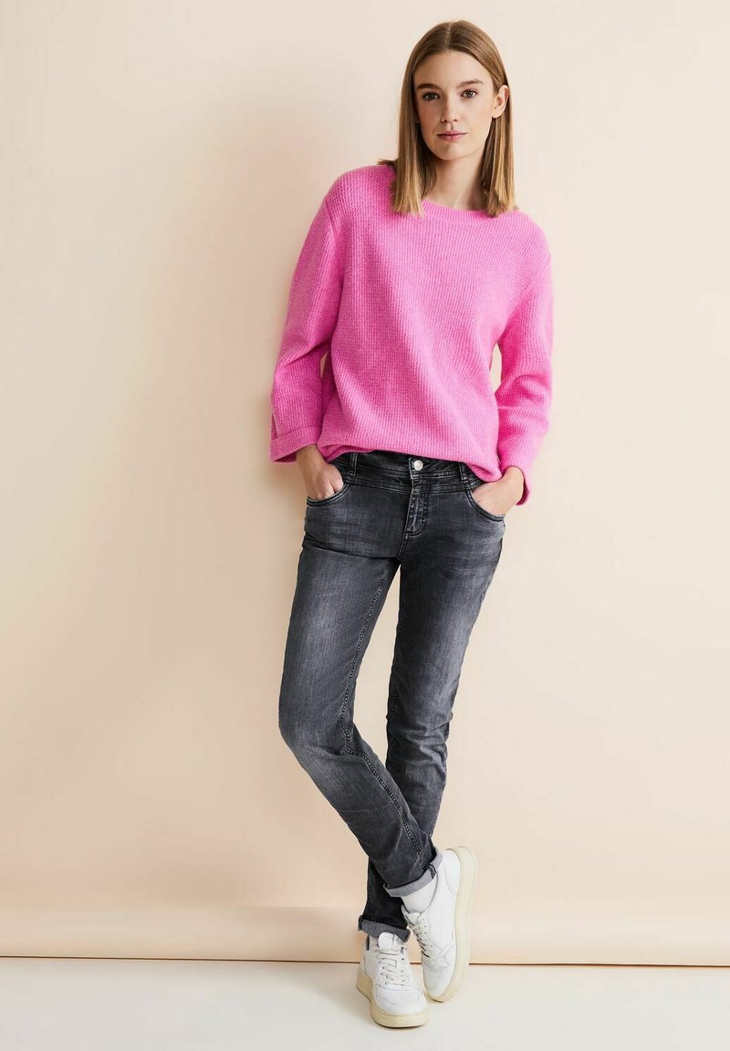 Suéter de punto rosa con patrón texturizado, de corte relajado, combinado con jeans gris oscuro y zapatillas blancas. Ambientado en un fondo neutral.