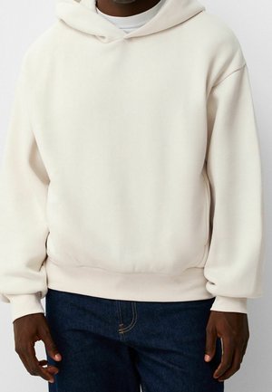 Mann trägt einen schlichten cremeweißen Kapuzenpullover und dunkelblaue Jeans, steht vor einem hellen Hintergrund.