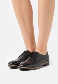 Chaussures en cuir noir à finition lisse, bout arrondi et petit talon empilé, avec un design à lacets et des accents marron subtils le long de la semelle.