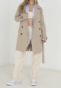 Beżowy płaszcz trench double-breasted z czarnymi guzikami, nałożony na jasnoniebieską koszulę w paski. W zestawieniu z kremowymi spodniami w szerokim kroju i białymi sneakersami.