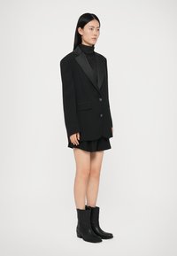 Zwarte blazer met een satijnen revers, gecombineerd met een zwarte coltrui, zwarte shorts en zwarte leren laarzen tot de knie. Soepele textuur overal.